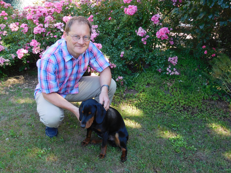 Dr. König kniet mit seinem Hund auf einer Wiese vor blühenden Büschen
