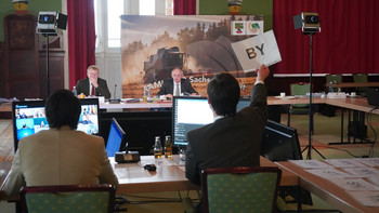 Blick auf einen Bildschirm, der die per Video zugeschalteten Konferenzteilnehmer zeigt. Dahinter ist Sachsen-Anhalts Amtschef Gert Zender vor Ort in Magdeburg zu sehen