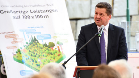 Forstminister Sven Schulze spricht beim Wirtschaftswaldgipfel in Schierke, im Hintergrund ist eine Grafik zu sehen, auf der die Leistung von 100 mal 100 Meter Wald zu erklärt werden.